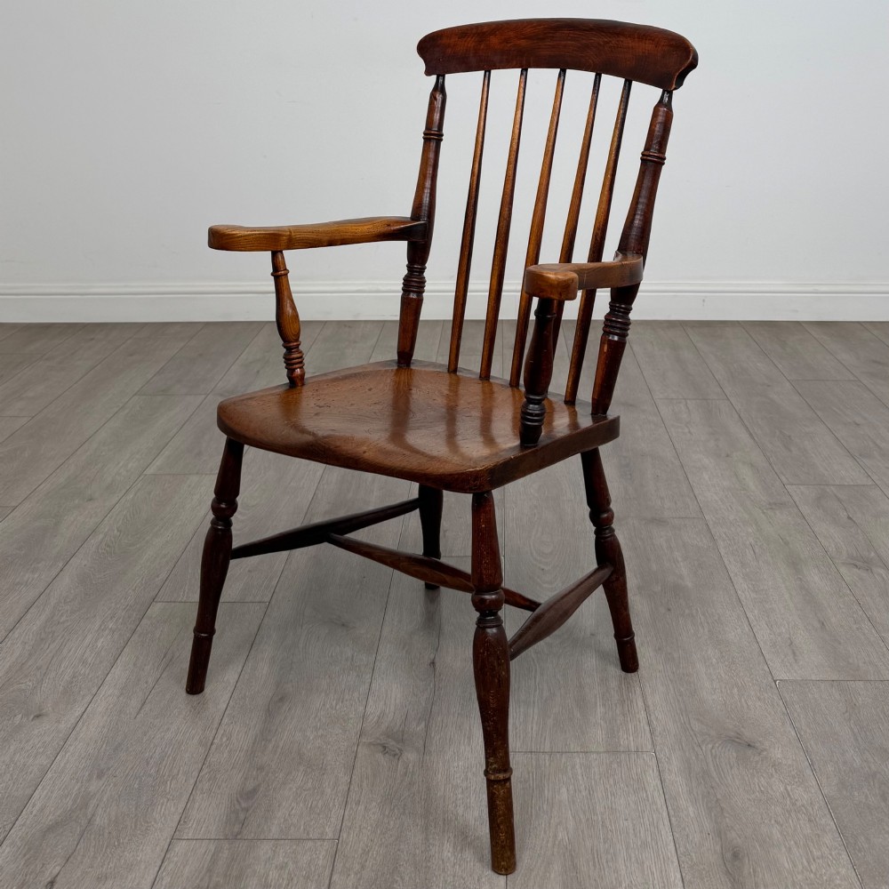 An antique 19th-century oak stick back armchair featuring turned spindle backrests, curved wooden armrests, a contoured solid seat, and turned legs joined by stretchers.