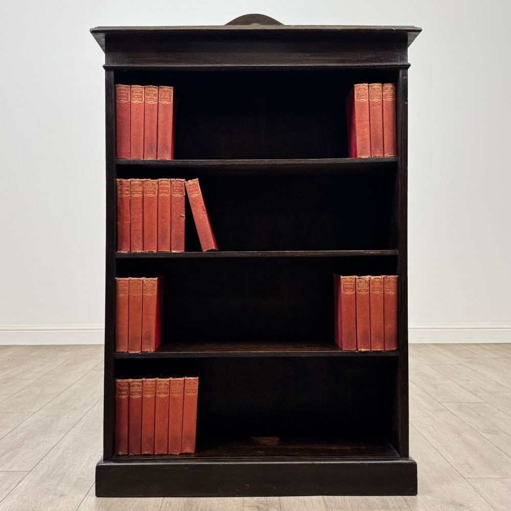 An Edwardian dark-stained oak open bookcase featuring four shelf tiers containing red leather-bound books against a white background.