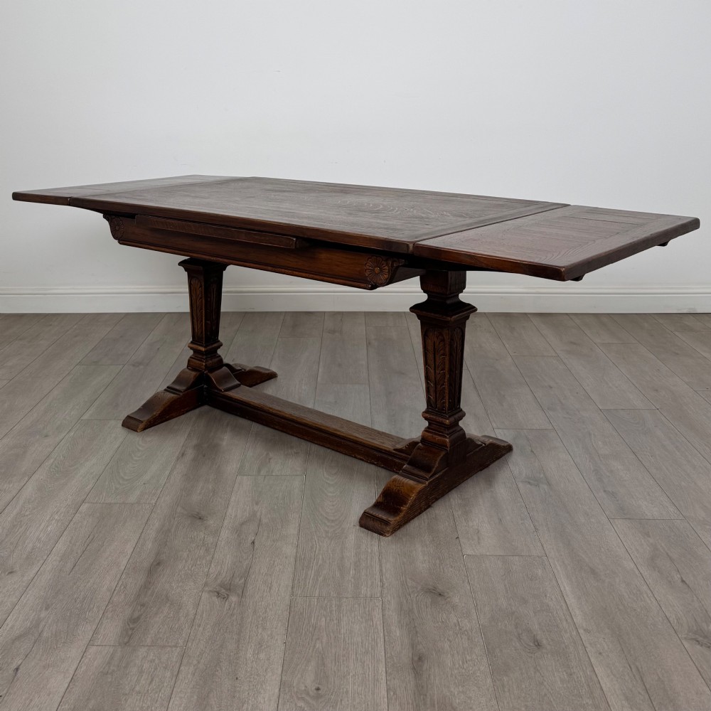 An Edwardian dark oak draw leaf dining table featuring extended side leaves, carved pedestal supports, and a low connecting stretcher.