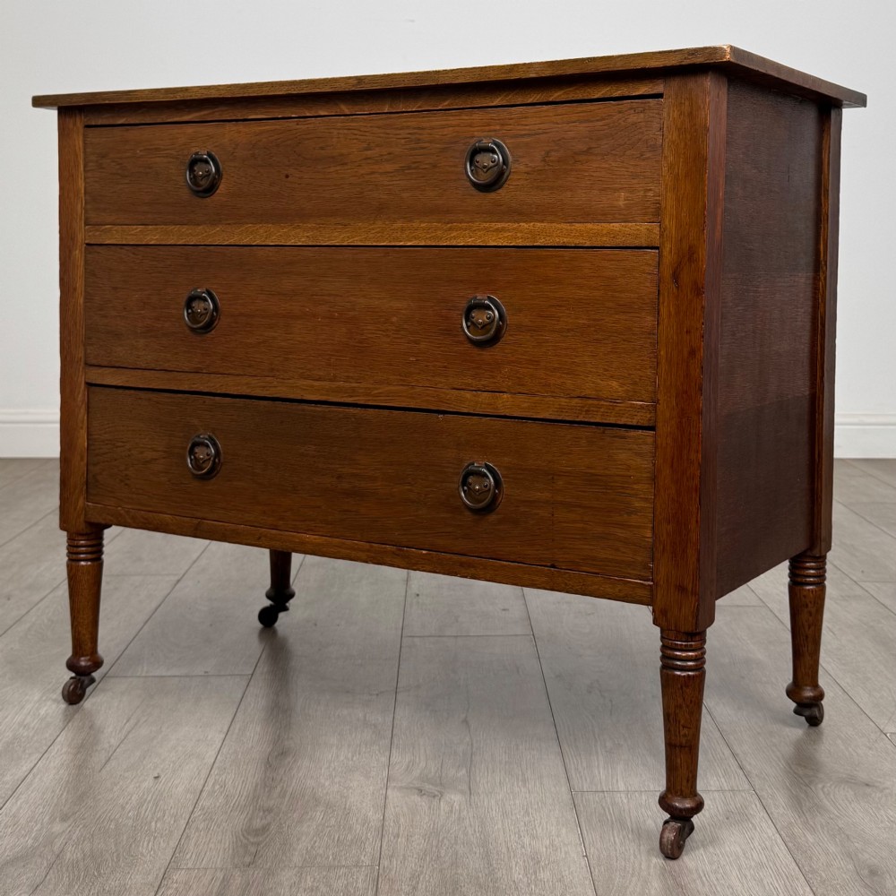 An antique Edwardian oak chest featuring three full-width drawers with brass ring drop handles, set on turned legs with castors. View II