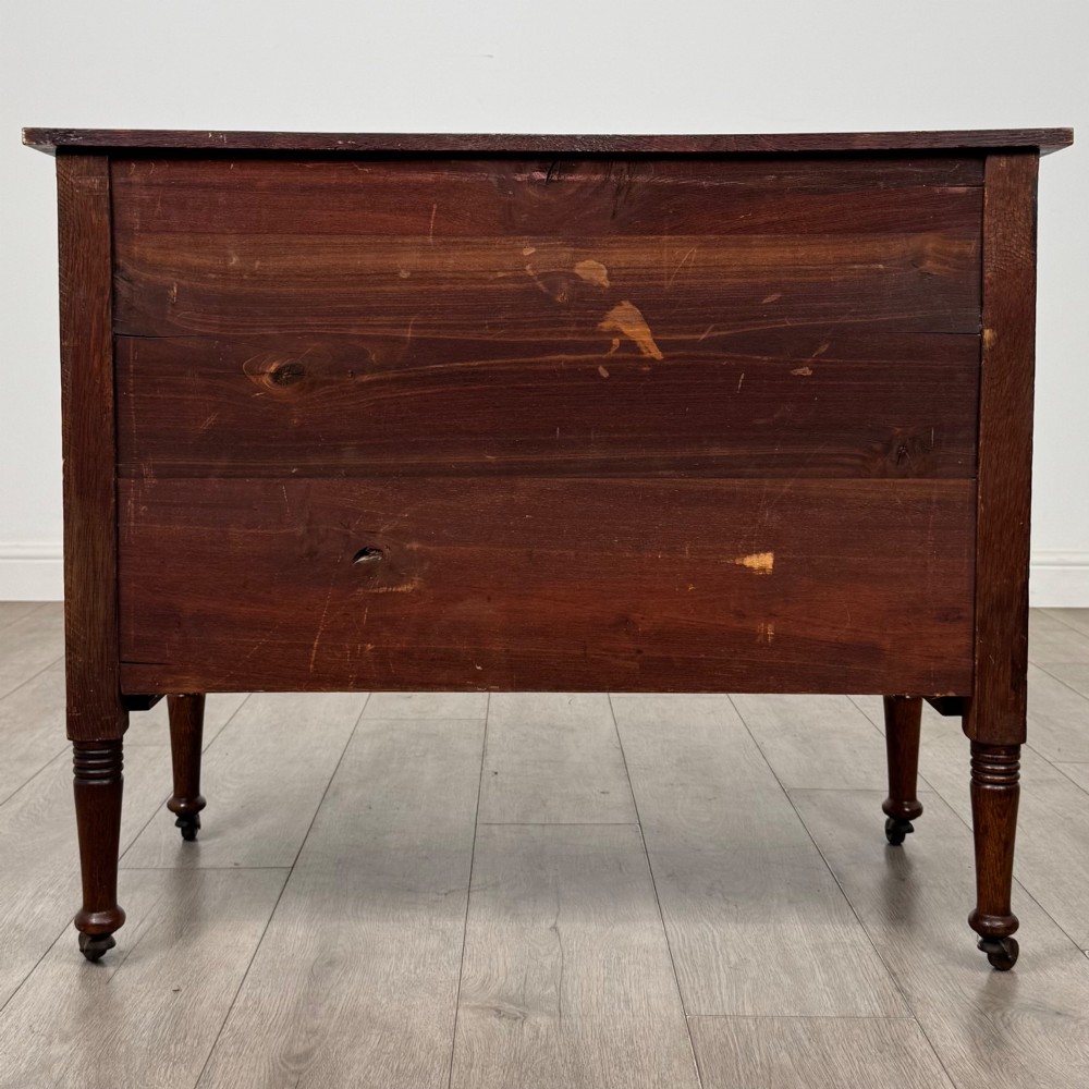 An antique Edwardian oak chest featuring three full-width drawers with brass ring drop handles, set on turned legs with castors. View V