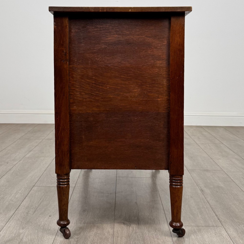 An antique Edwardian oak chest featuring three full-width drawers with brass ring drop handles, set on turned legs with castors. View VII