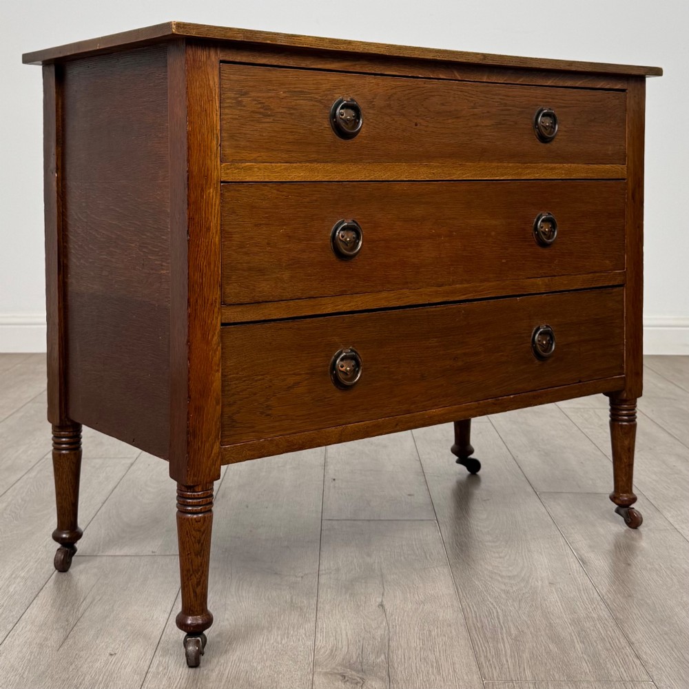 An antique Edwardian oak chest featuring three full-width drawers with brass ring drop handles, set on turned legs with castors. View VIII