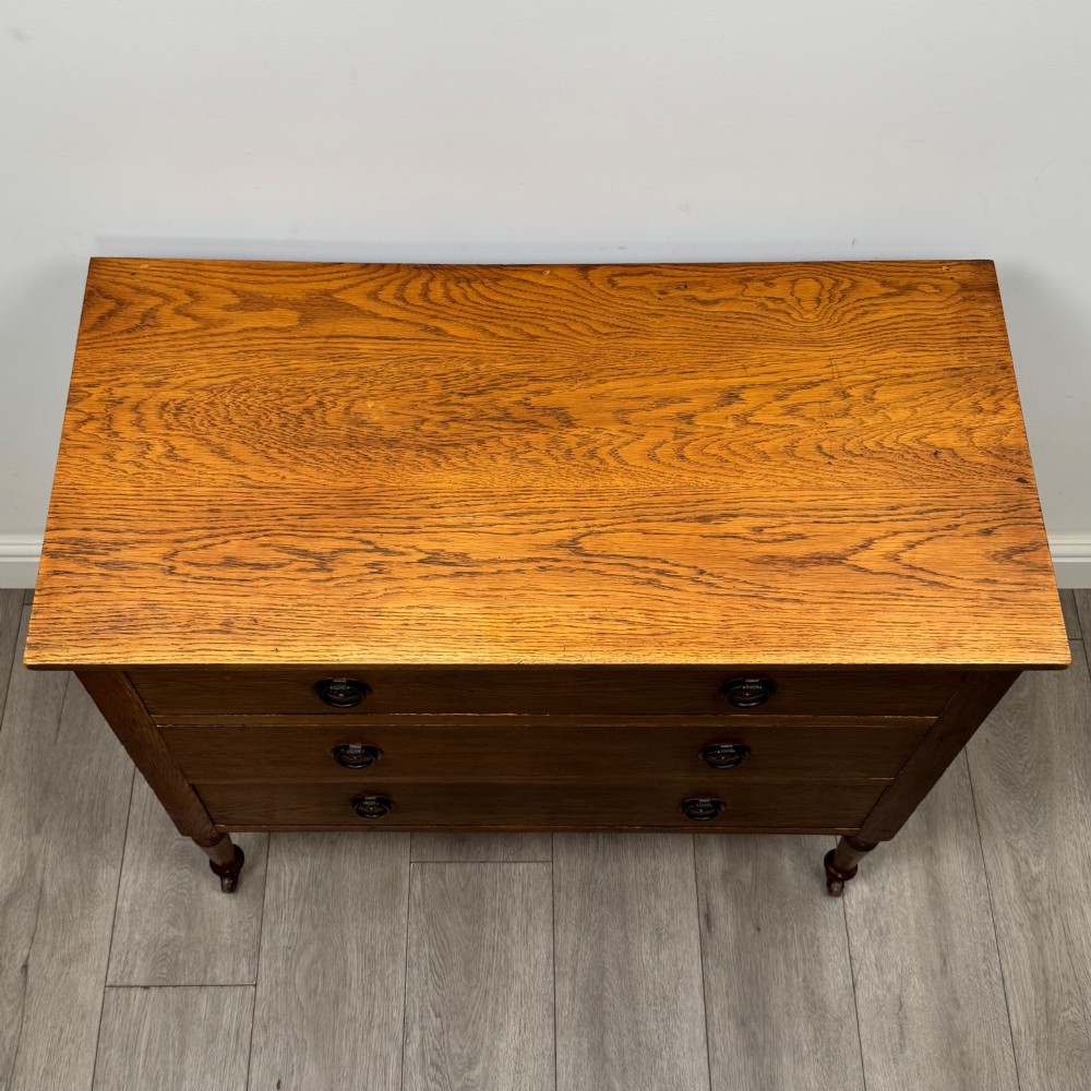 An antique Edwardian oak chest featuring three full-width drawers with brass ring drop handles, set on turned legs with castors. View IX