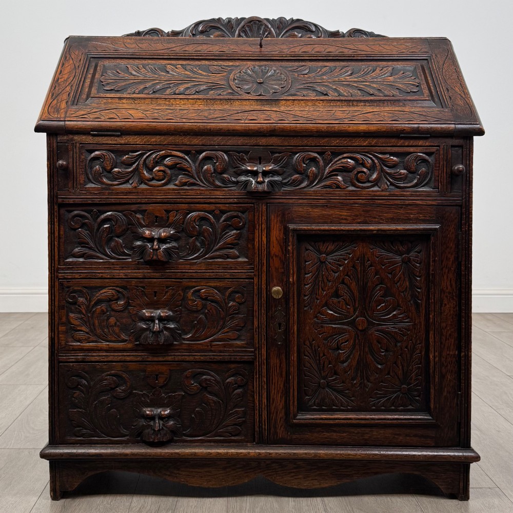 A dark oak Victorian slant-front bureau featuring carved Green Man drawer pulls, foliage relief carvings, and a single panelled cupboard door.