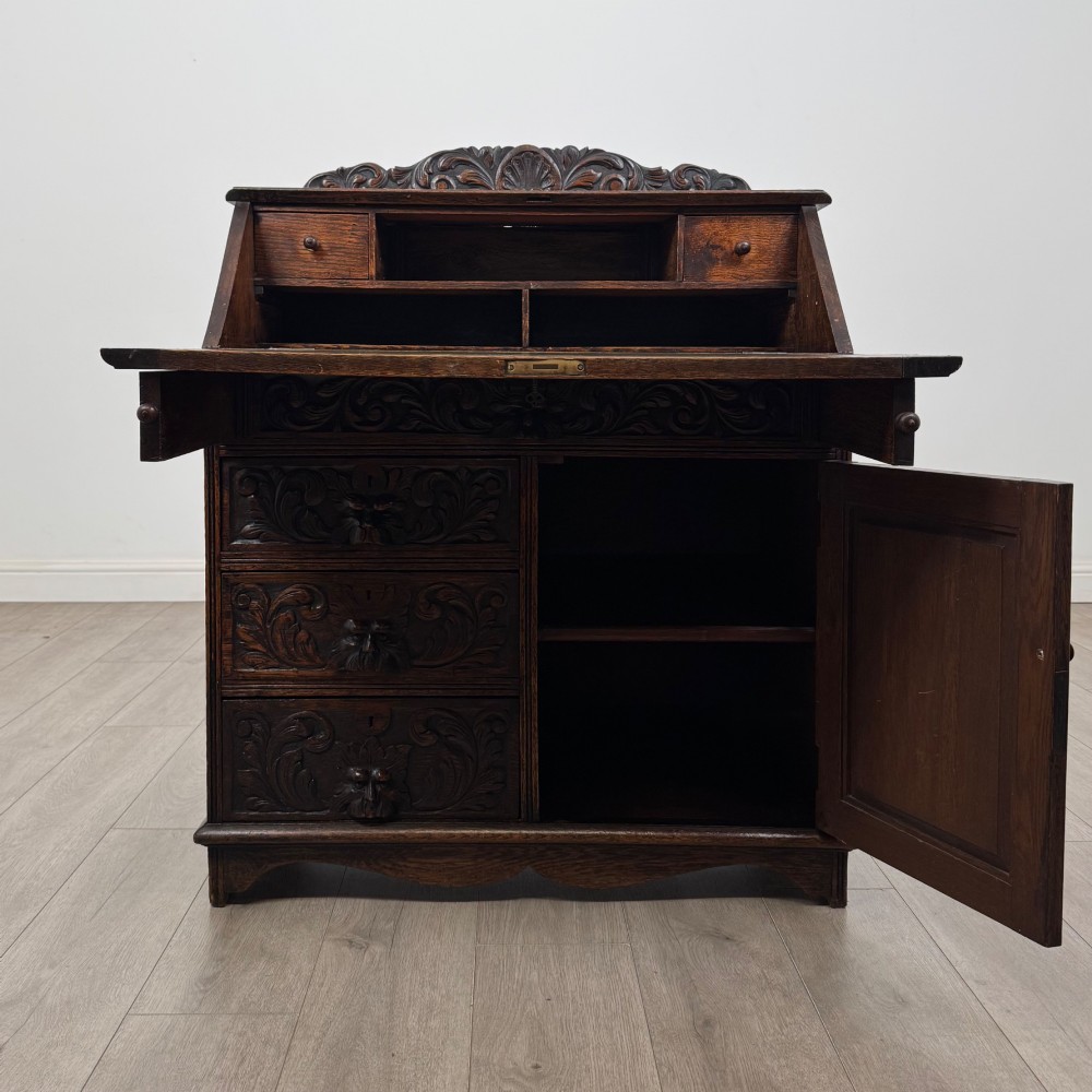 A dark oak Victorian slant-front bureau featuring carved Green Man drawer pulls, foliage relief carvings, and a single panelled cupboard door. View II