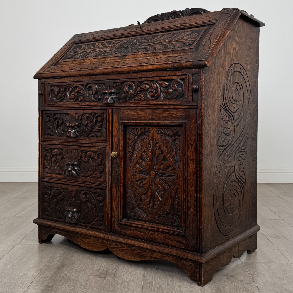 A dark oak Victorian slant-front bureau featuring carved Green Man drawer pulls, foliage relief carvings, and a single panelled cupboard door. View III