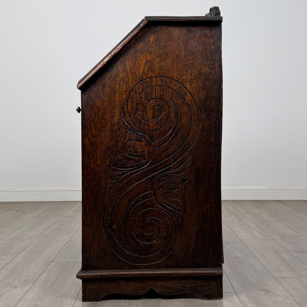 A dark oak Victorian slant-front bureau featuring carved Green Man drawer pulls, foliage relief carvings, and a single panelled cupboard door. View IV