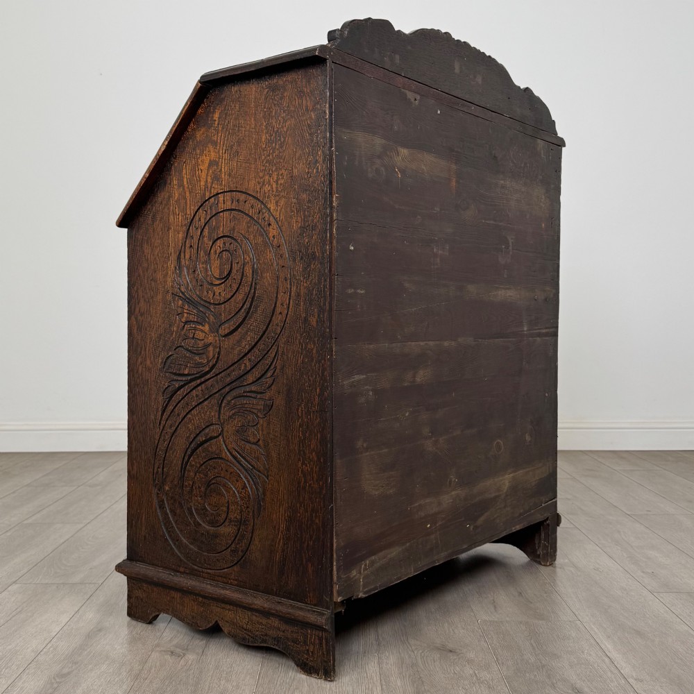 A dark oak Victorian slant-front bureau featuring carved Green Man drawer pulls, foliage relief carvings, and a single panelled cupboard door. View V