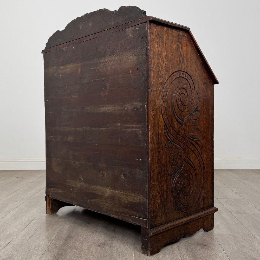 A dark oak Victorian slant-front bureau featuring carved Green Man drawer pulls, foliage relief carvings, and a single panelled cupboard door. View VII