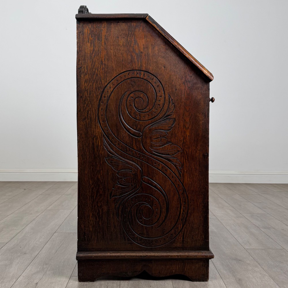 A dark oak Victorian slant-front bureau featuring carved Green Man drawer pulls, foliage relief carvings, and a single panelled cupboard door. View VIII