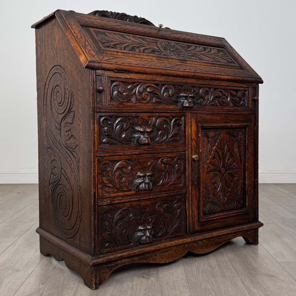 A dark oak Victorian slant-front bureau featuring carved Green Man drawer pulls, foliage relief carvings, and a single panelled cupboard door. View IX