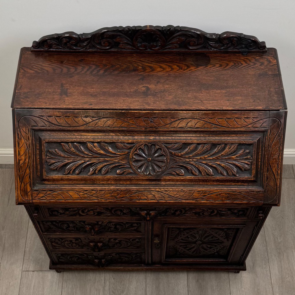 A dark oak Victorian slant-front bureau featuring carved Green Man drawer pulls, foliage relief carvings, and a single panelled cupboard door. View X