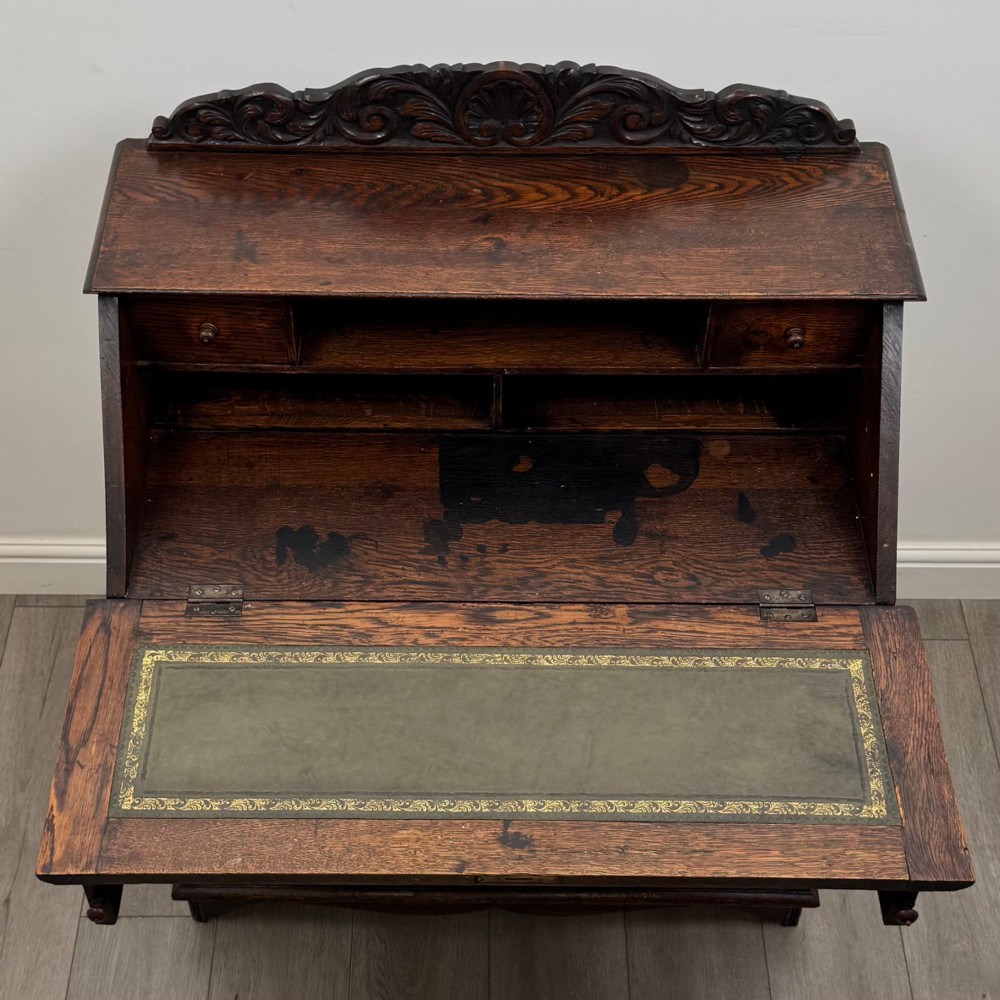 A dark oak Victorian slant-front bureau featuring carved Green Man drawer pulls, foliage relief carvings, and a single panelled cupboard door. View XI