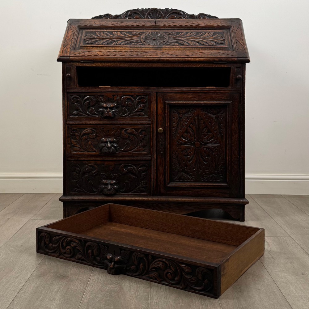 A dark oak Victorian slant-front bureau featuring carved Green Man drawer pulls, foliage relief carvings, and a single panelled cupboard door. View XII