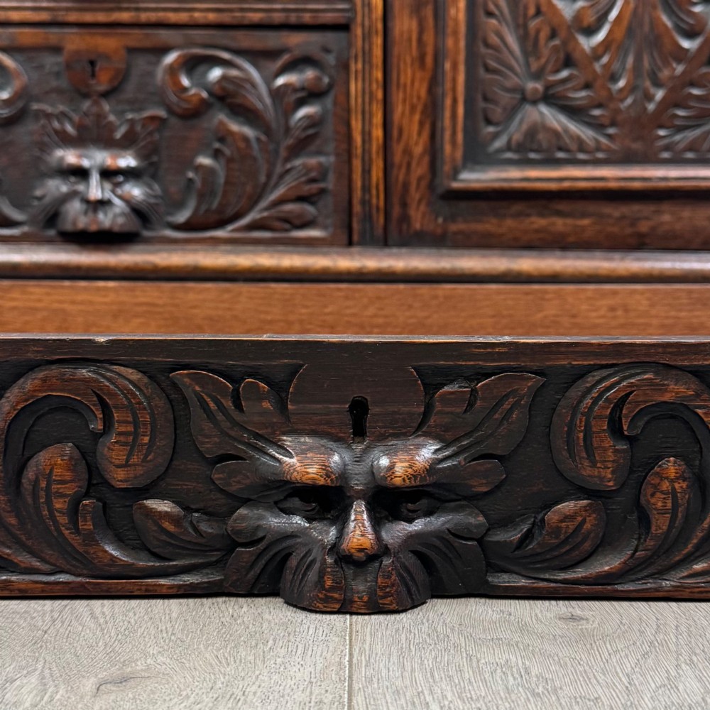 A dark oak Victorian slant-front bureau featuring carved Green Man drawer pulls, foliage relief carvings, and a single panelled cupboard door. View XIII