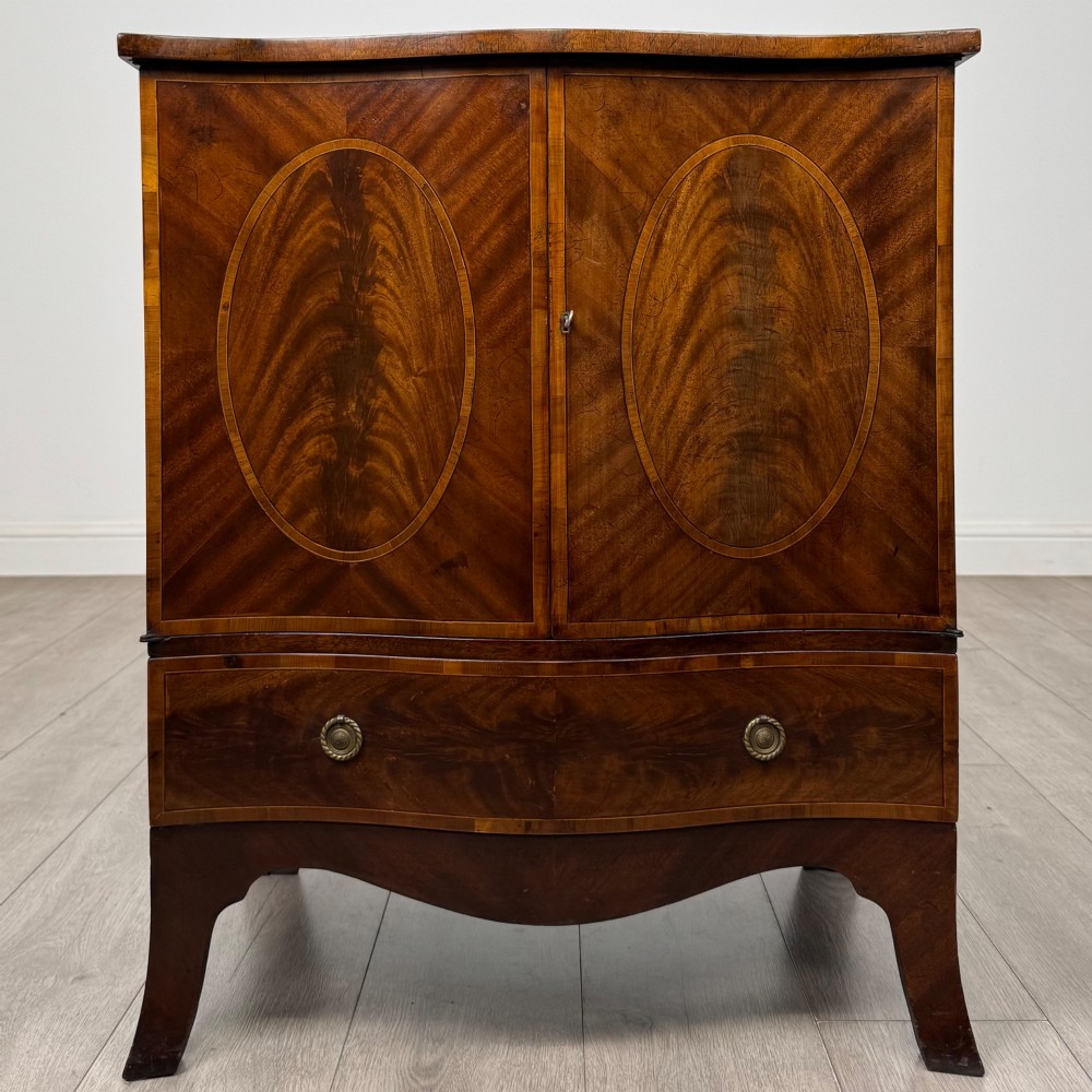 A 20th-century mahogany serpentine cupboard featuring flame mahogany oval door panels, chevron veneering, a lower drawer with brass ring handles, and splayed bracket feet.