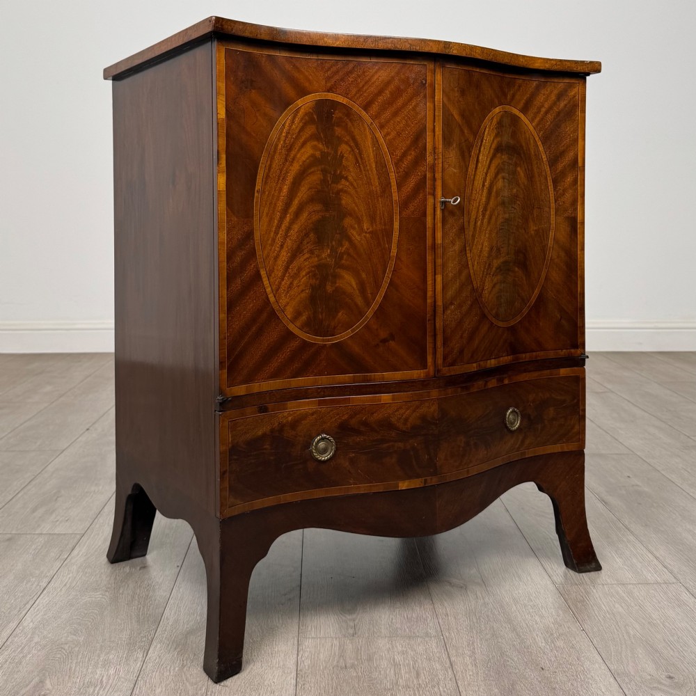 A 20th-century mahogany serpentine cupboard featuring flame mahogany oval door panels, chevron veneering, a lower drawer with brass ring handles, and splayed bracket feet. View IX