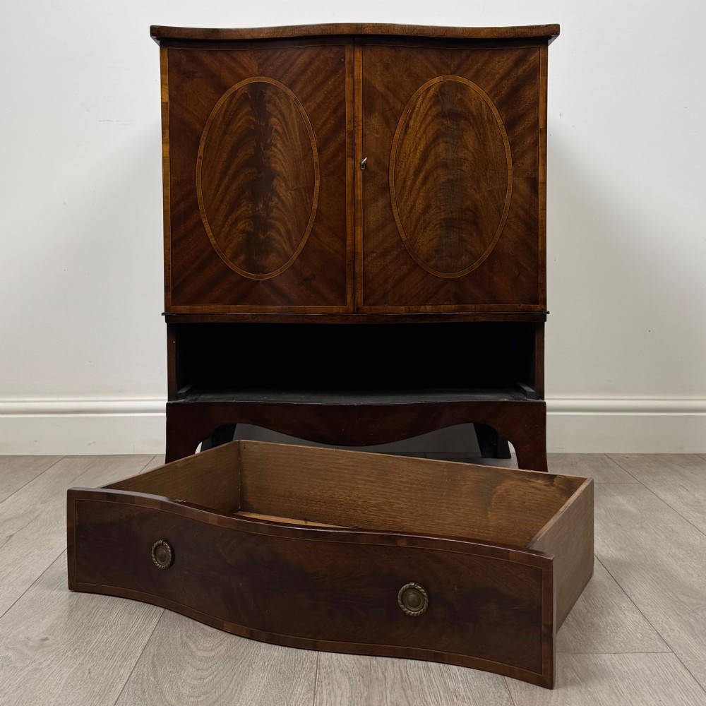 A 20th-century mahogany serpentine cupboard featuring flame mahogany oval door panels, chevron veneering, a lower drawer with brass ring handles, and splayed bracket feet. View XI
