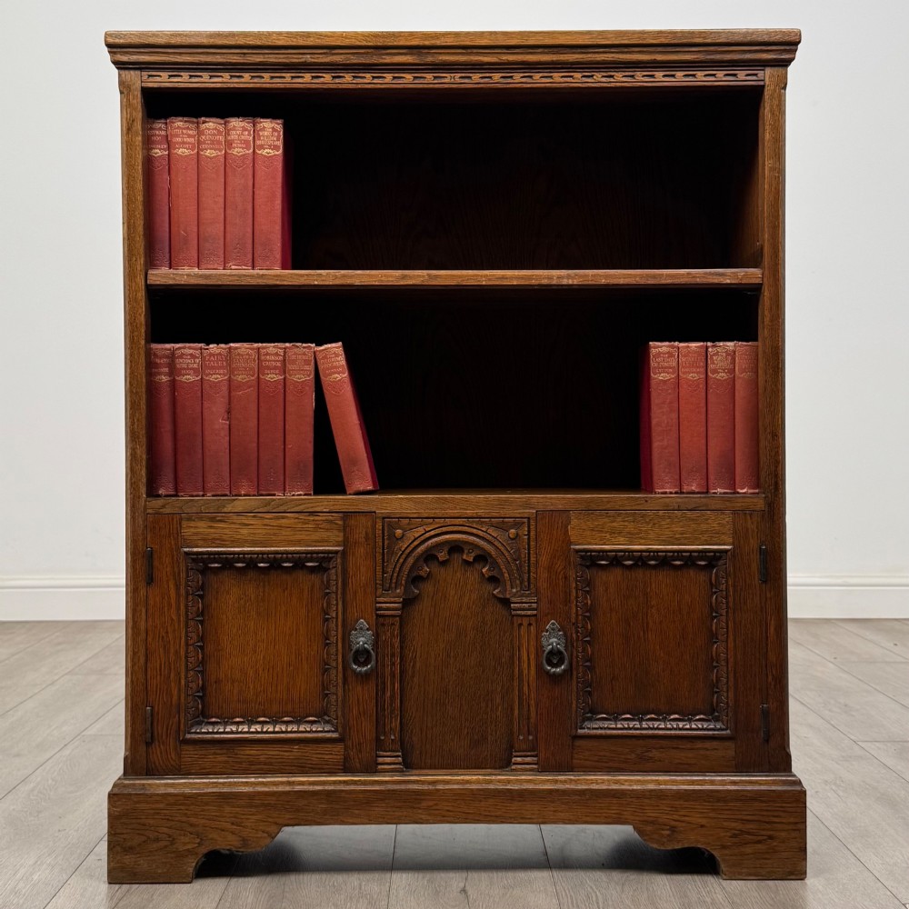 A small medium-brown oak open bookcase featuring two upper shelves and a lower two-door cupboard with carved arched motifs and metal ring handles.