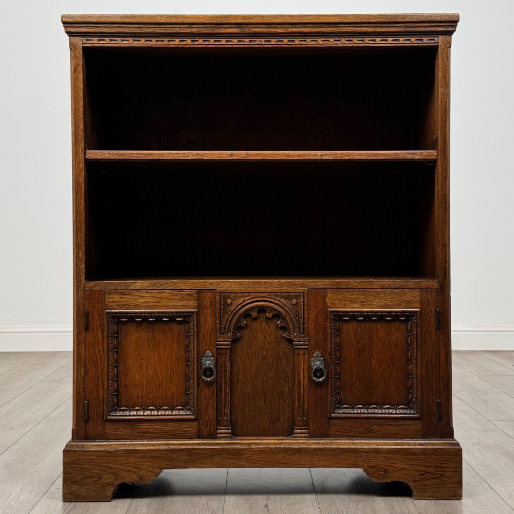 A small medium-brown oak open bookcase featuring two upper shelves and a lower two-door cupboard with carved arched motifs and metal ring handles. View II