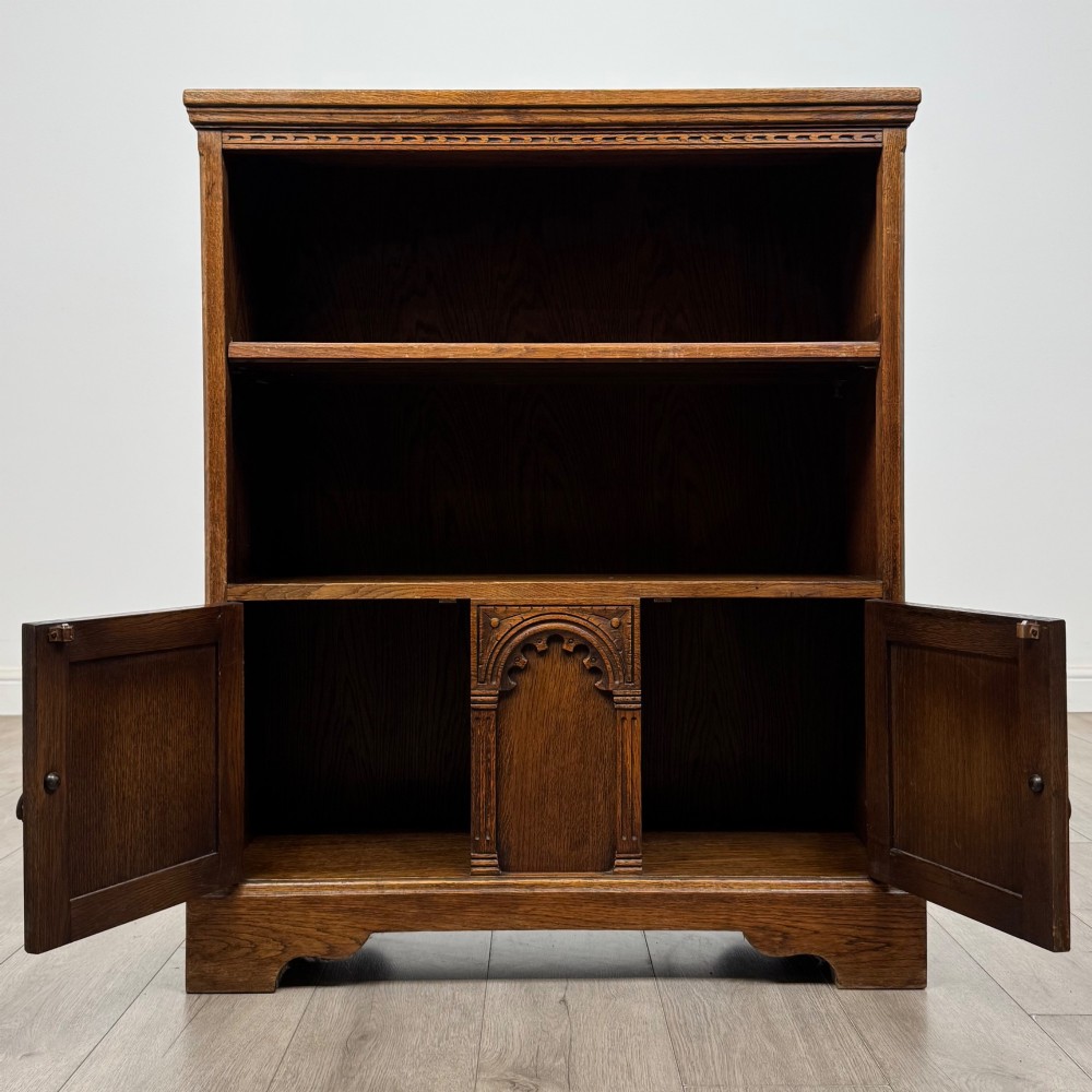 A small medium-brown oak open bookcase featuring two upper shelves and a lower two-door cupboard with carved arched motifs and metal ring handles. View III