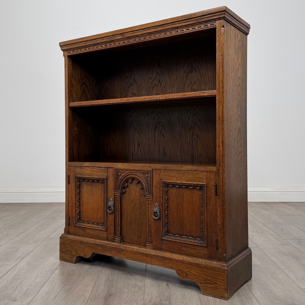 A small medium-brown oak open bookcase featuring two upper shelves and a lower two-door cupboard with carved arched motifs and metal ring handles. View IV