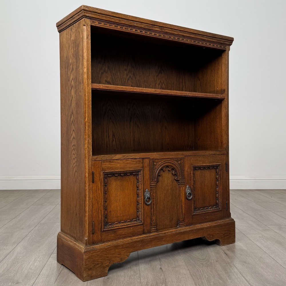A small medium-brown oak open bookcase featuring two upper shelves and a lower two-door cupboard with carved arched motifs and metal ring handles. View X