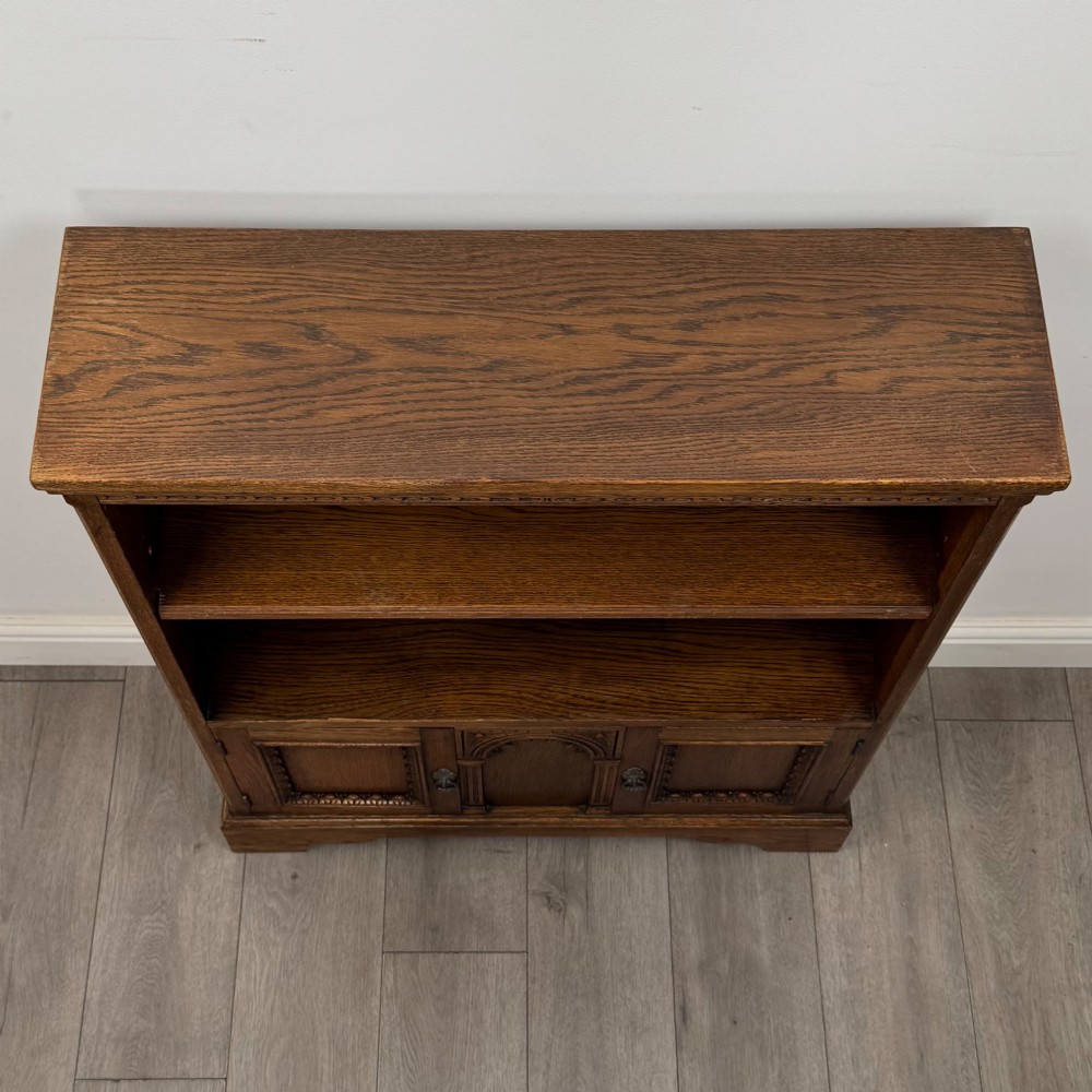 A small medium-brown oak open bookcase featuring two upper shelves and a lower two-door cupboard with carved arched motifs and metal ring handles. View XI