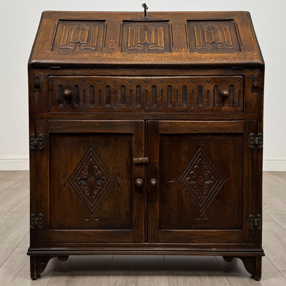 A dark oak slant-front writing bureau featuring carved linenfold upper panels, a single carved drawer with wooden knobs, and a double-door cupboard base decorated with diamond lozenge carvings.