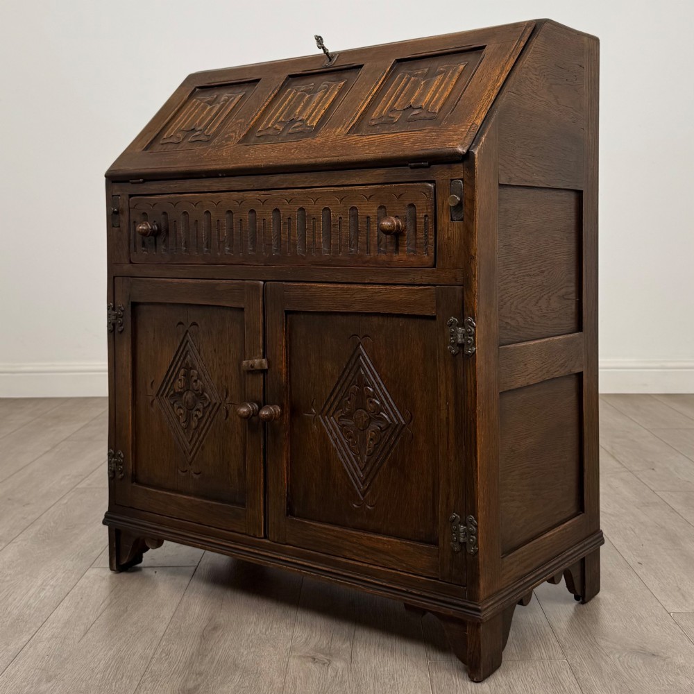 A dark oak slant-front writing bureau featuring carved linenfold upper panels, a single carved drawer with wooden knobs, and a double-door cupboard base decorated with diamond lozenge carvings. View III