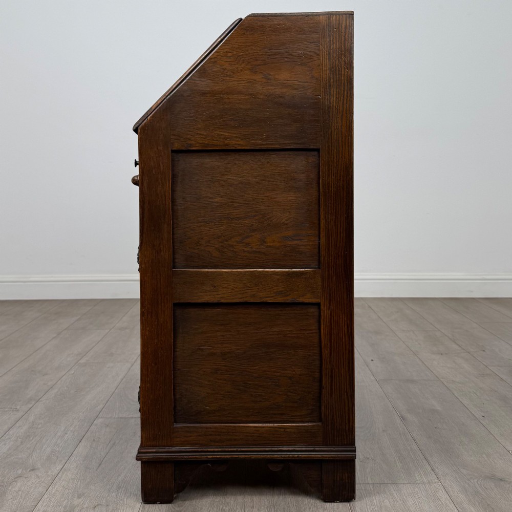 A dark oak slant-front writing bureau featuring carved linenfold upper panels, a single carved drawer with wooden knobs, and a double-door cupboard base decorated with diamond lozenge carvings. View IV
