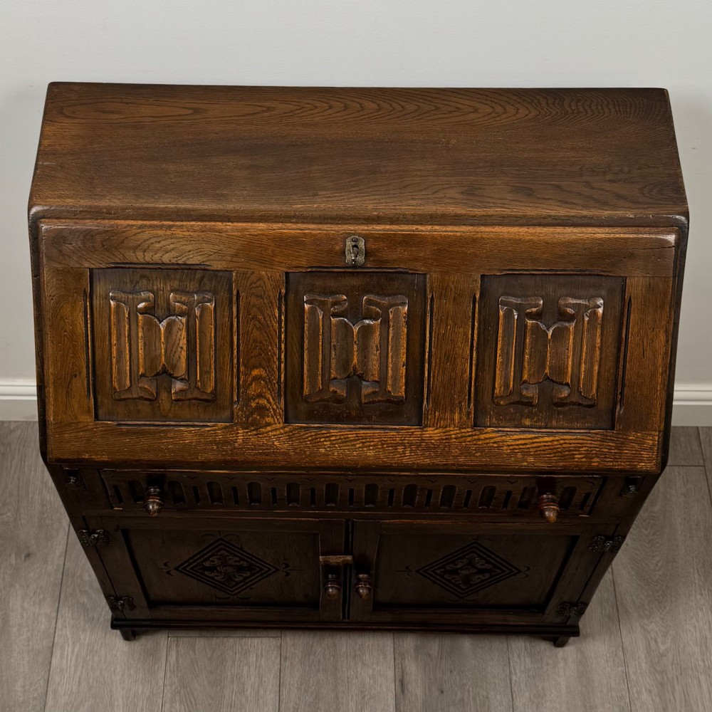 A dark oak slant-front writing bureau featuring carved linenfold upper panels, a single carved drawer with wooden knobs, and a double-door cupboard base decorated with diamond lozenge carvings. View X