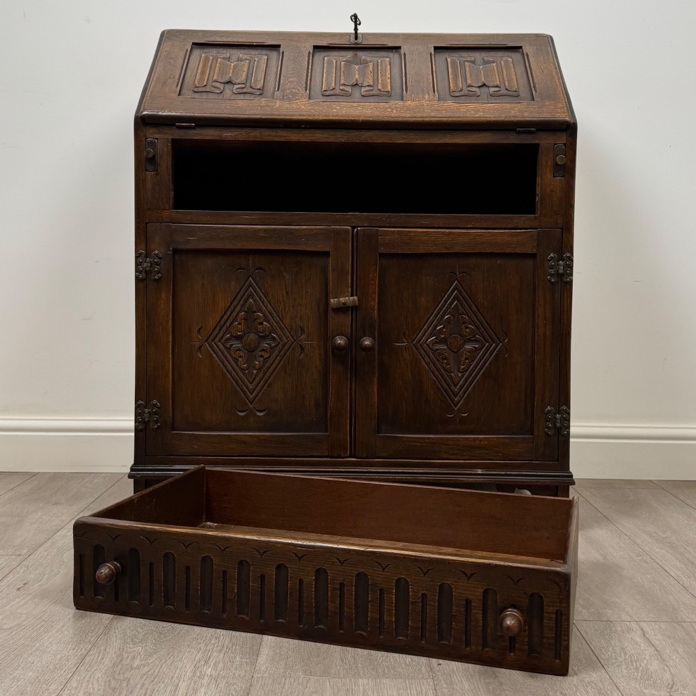 A dark oak slant-front writing bureau featuring carved linenfold upper panels, a single carved drawer with wooden knobs, and a double-door cupboard base decorated with diamond lozenge carvings. View XII