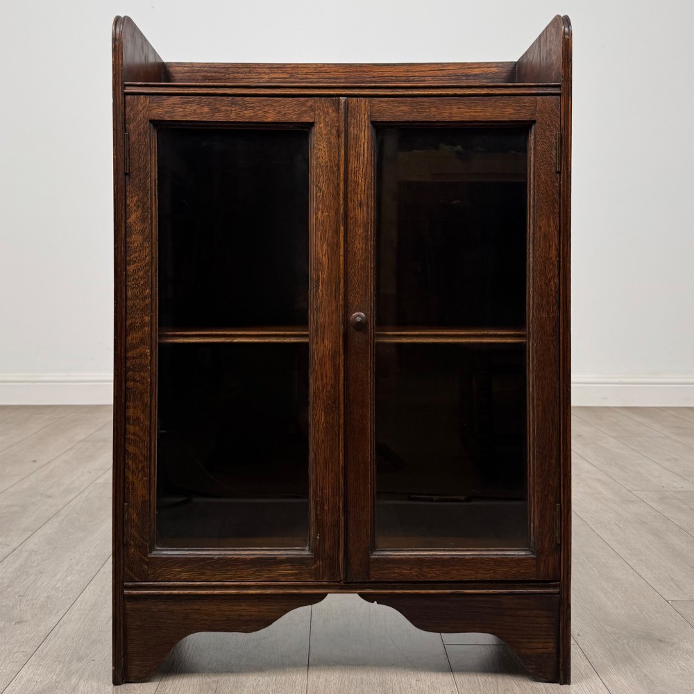 An early 20th-century dark oak display cabinet featuring twin glazed doors, an interior shelf, and a gallery top.