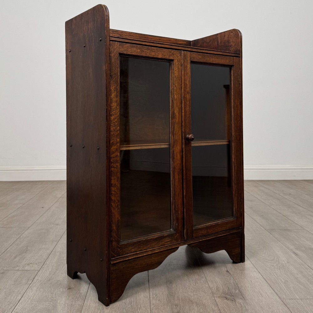 An early 20th-century dark oak display cabinet featuring twin glazed doors, an interior shelf, and a gallery top. View IX