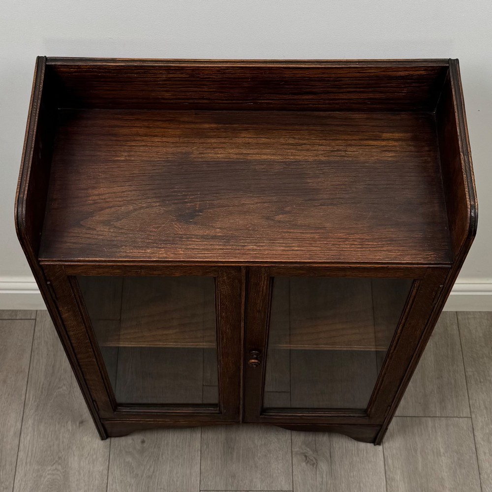 An early 20th-century dark oak display cabinet featuring twin glazed doors, an interior shelf, and a gallery top. View X