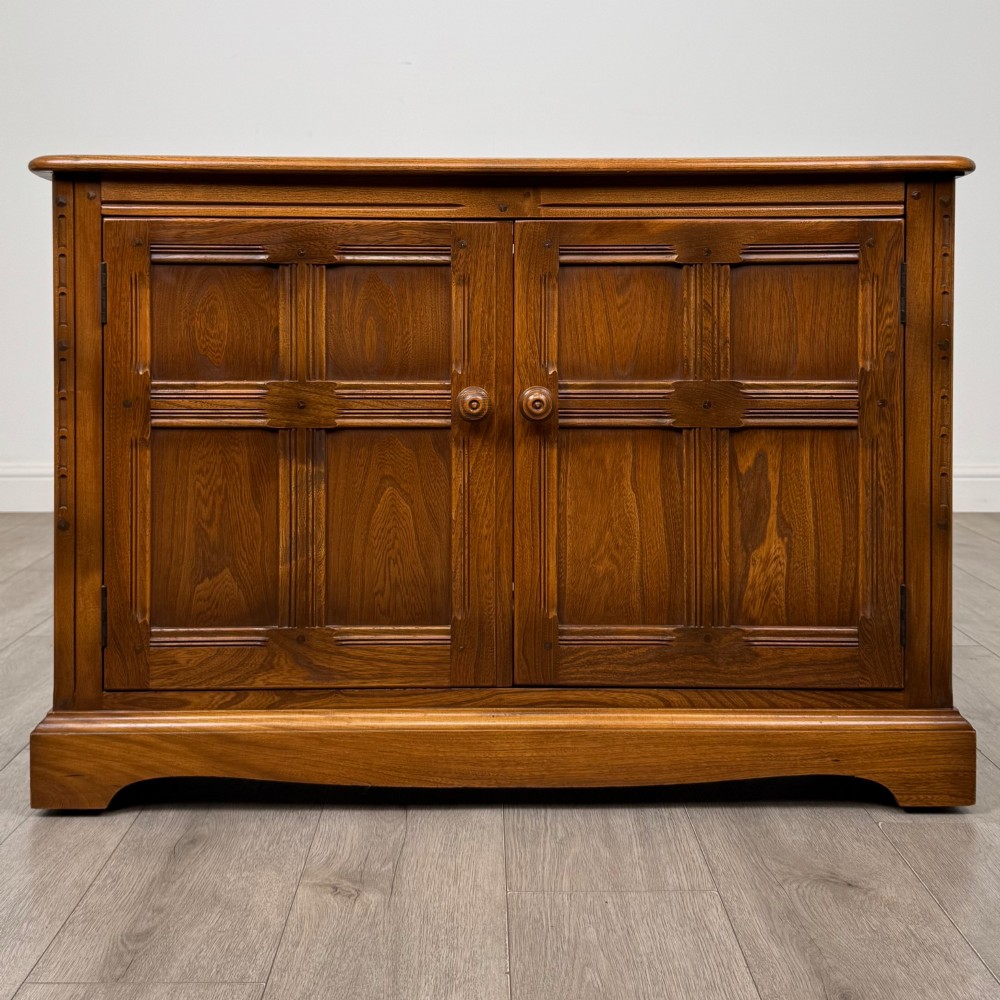 A two-door golden oak storage cupboard featuring geometric panelled doors, turned timber knobs, and a low plinth base.