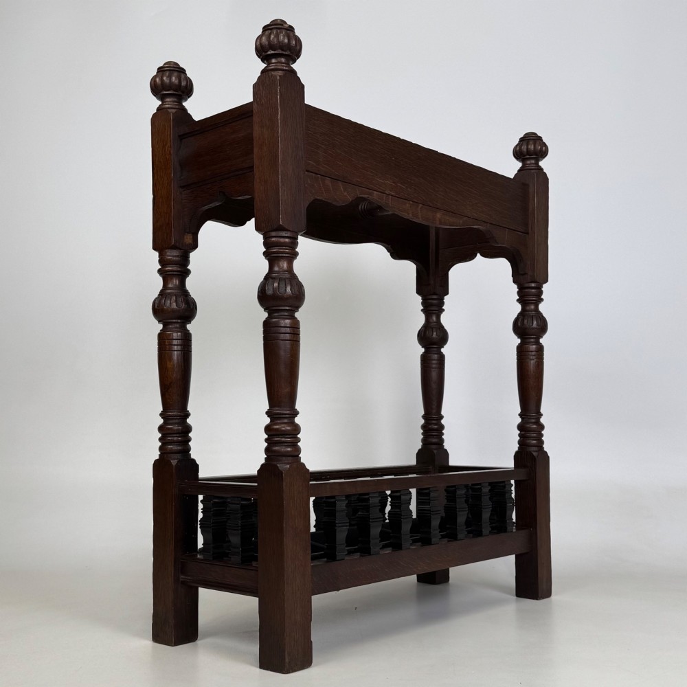 An Edwardian dark oak umbrella stand featuring four turned corner posts topped with finials, a shaped upper crossbar, and a lower spindle gallery enclosing a drip tray. View IV