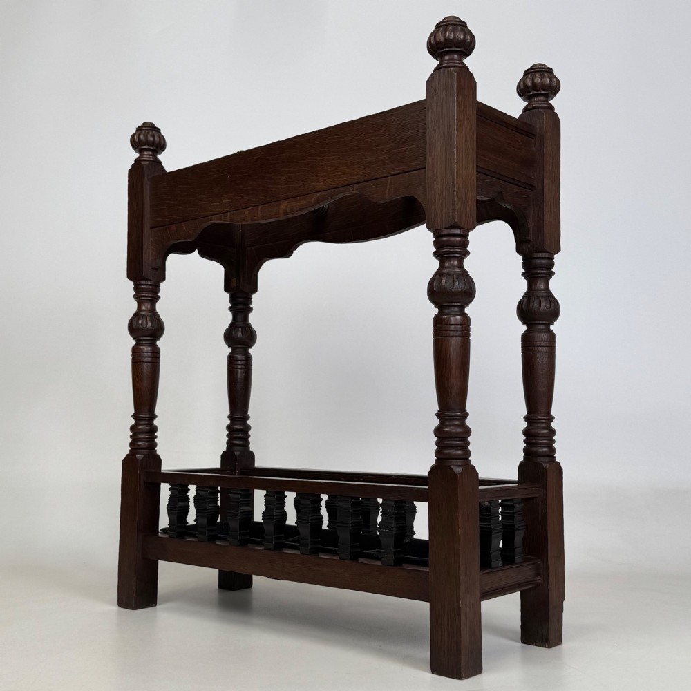 An Edwardian dark oak umbrella stand featuring four turned corner posts topped with finials, a shaped upper crossbar, and a lower spindle gallery enclosing a drip tray. View VI