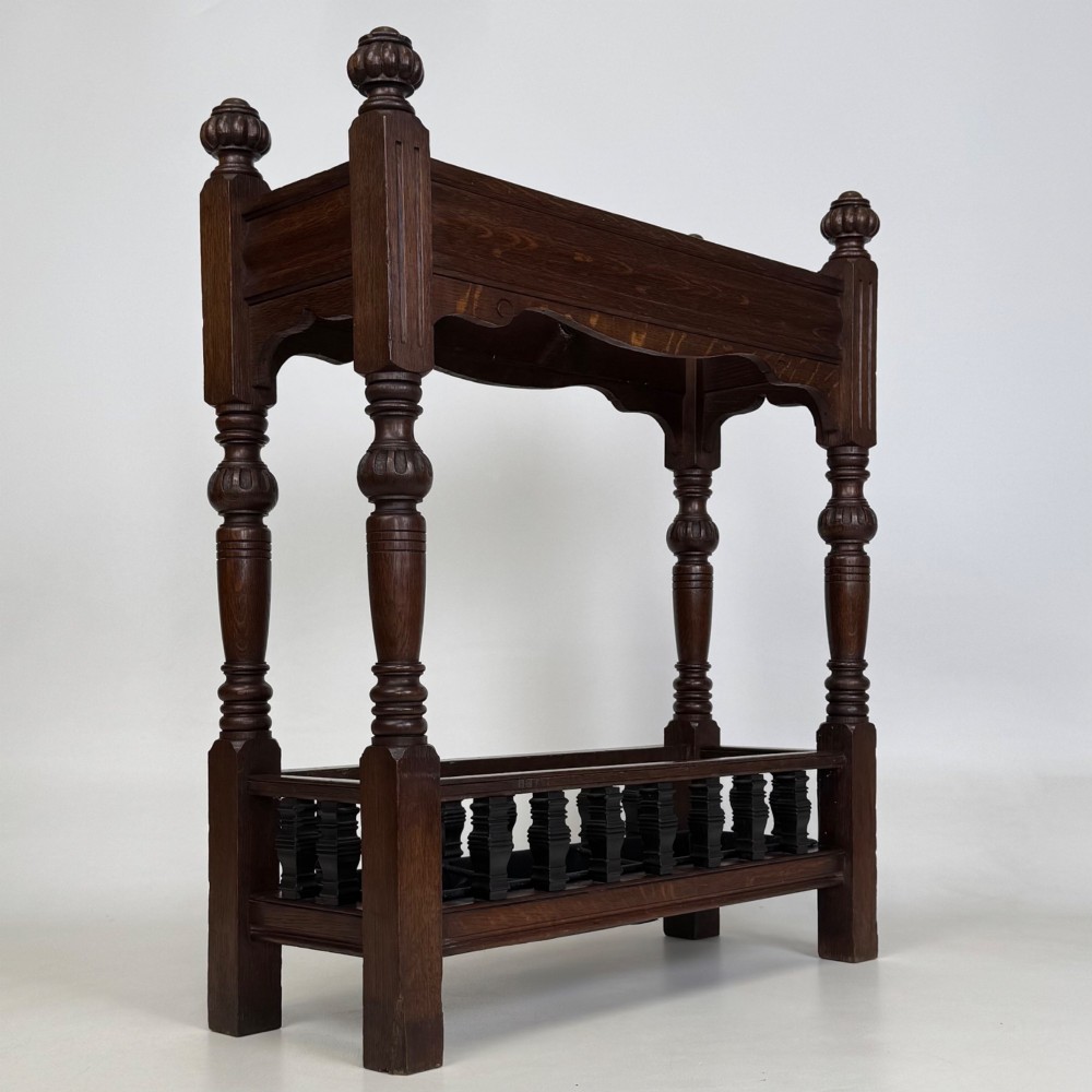 An Edwardian dark oak umbrella stand featuring four turned corner posts topped with finials, a shaped upper crossbar, and a lower spindle gallery enclosing a drip tray. View VIII