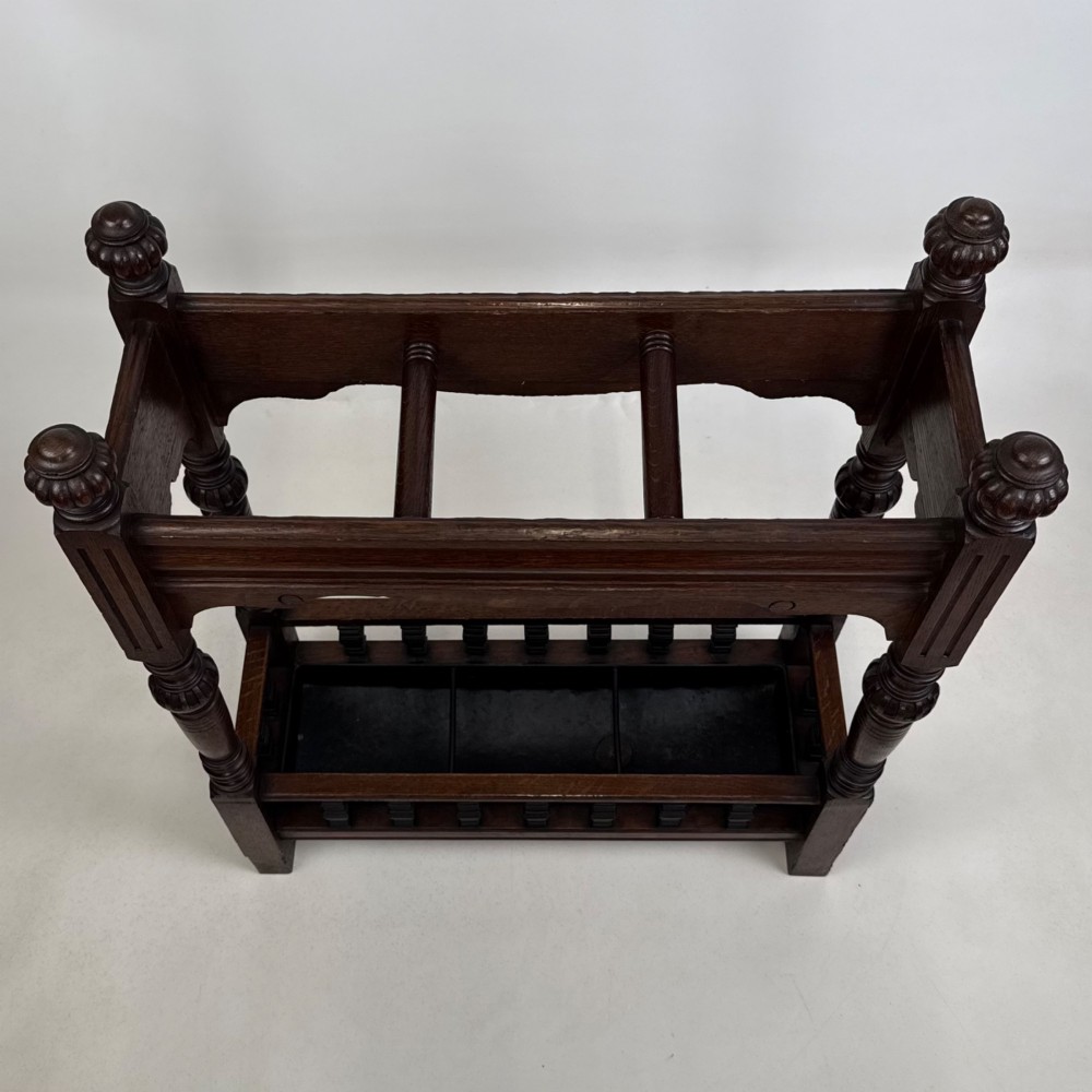 An Edwardian dark oak umbrella stand featuring four turned corner posts topped with finials, a shaped upper crossbar, and a lower spindle gallery enclosing a drip tray. View IX