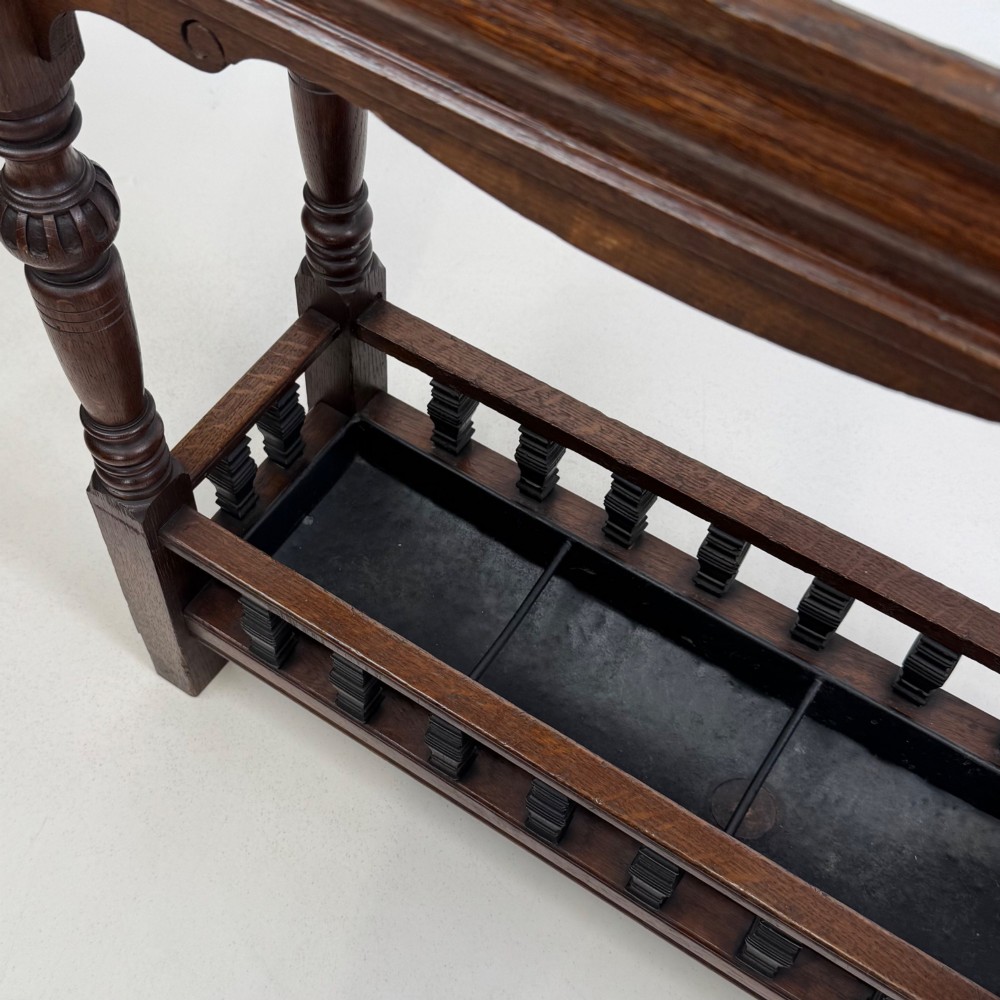 An Edwardian dark oak umbrella stand featuring four turned corner posts topped with finials, a shaped upper crossbar, and a lower spindle gallery enclosing a drip tray. View X