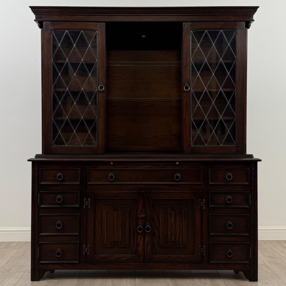 A dark oak display cabinet with a leaded glass central upper cupboard, flanked by open shelves, set atop a lower unit with multiple drawers and carved cupboard doors. View II