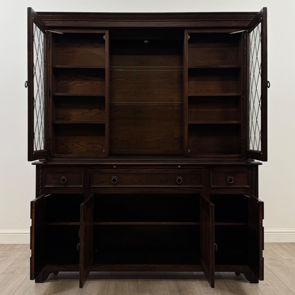 A dark oak display cabinet with a leaded glass central upper cupboard, flanked by open shelves, set atop a lower unit with multiple drawers and carved cupboard doors. View III