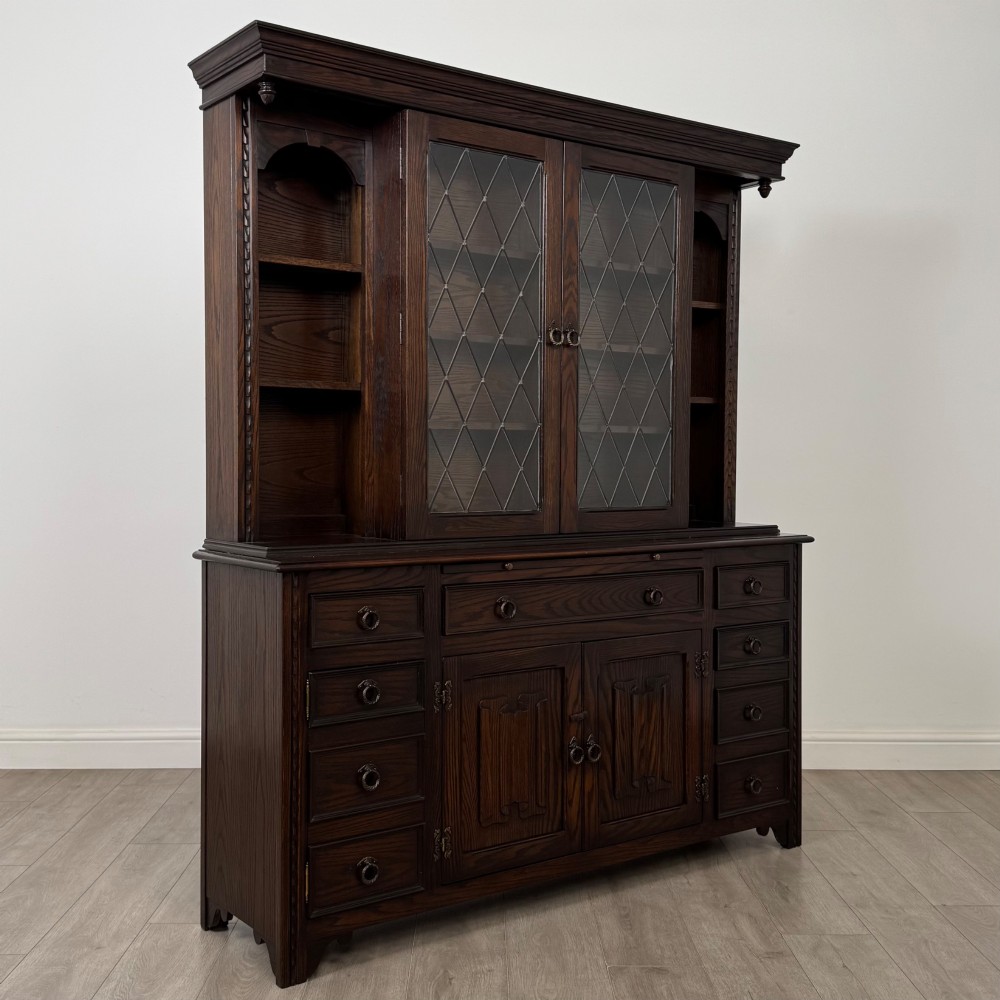 A dark oak display cabinet with a leaded glass central upper cupboard, flanked by open shelves, set atop a lower unit with multiple drawers and carved cupboard doors. View X
