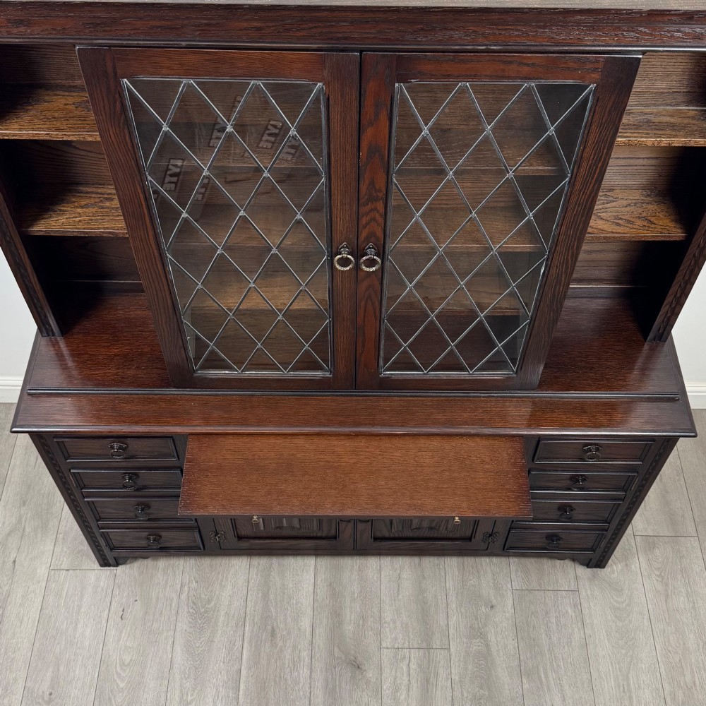A dark oak display cabinet with a leaded glass central upper cupboard, flanked by open shelves, set atop a lower unit with multiple drawers and carved cupboard doors. View XI