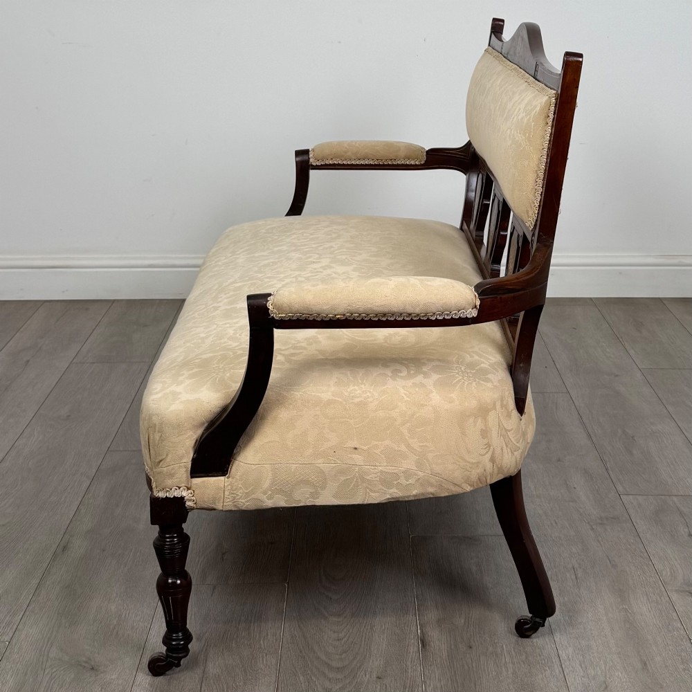 An antique Victorian rosewood settee featuring central backrest marquetry inlay, open armrests, turned front legs on castors, and cream damask upholstery. View III