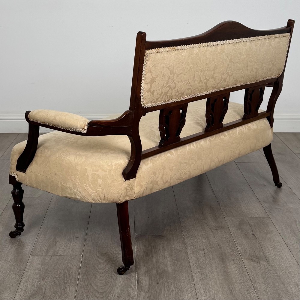 An antique Victorian rosewood settee featuring central backrest marquetry inlay, open armrests, turned front legs on castors, and cream damask upholstery. View IV