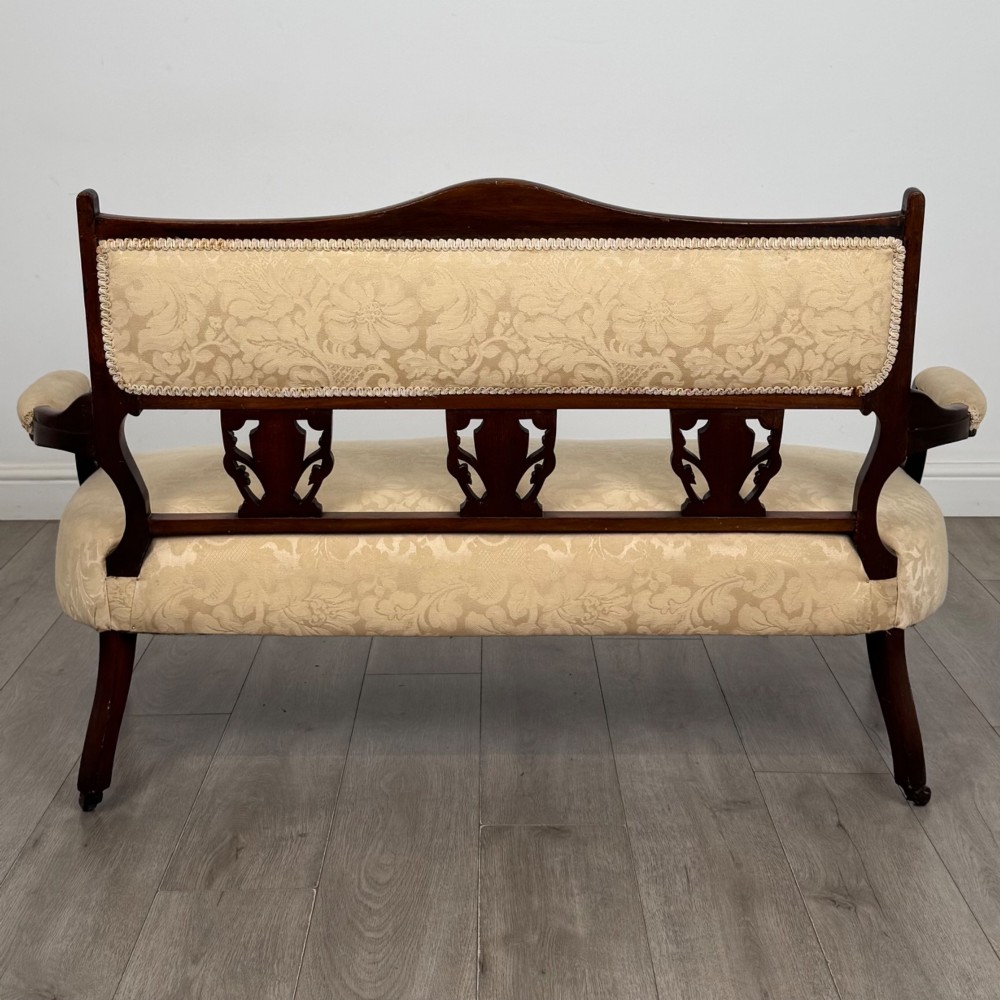 An antique Victorian rosewood settee featuring central backrest marquetry inlay, open armrests, turned front legs on castors, and cream damask upholstery. View V